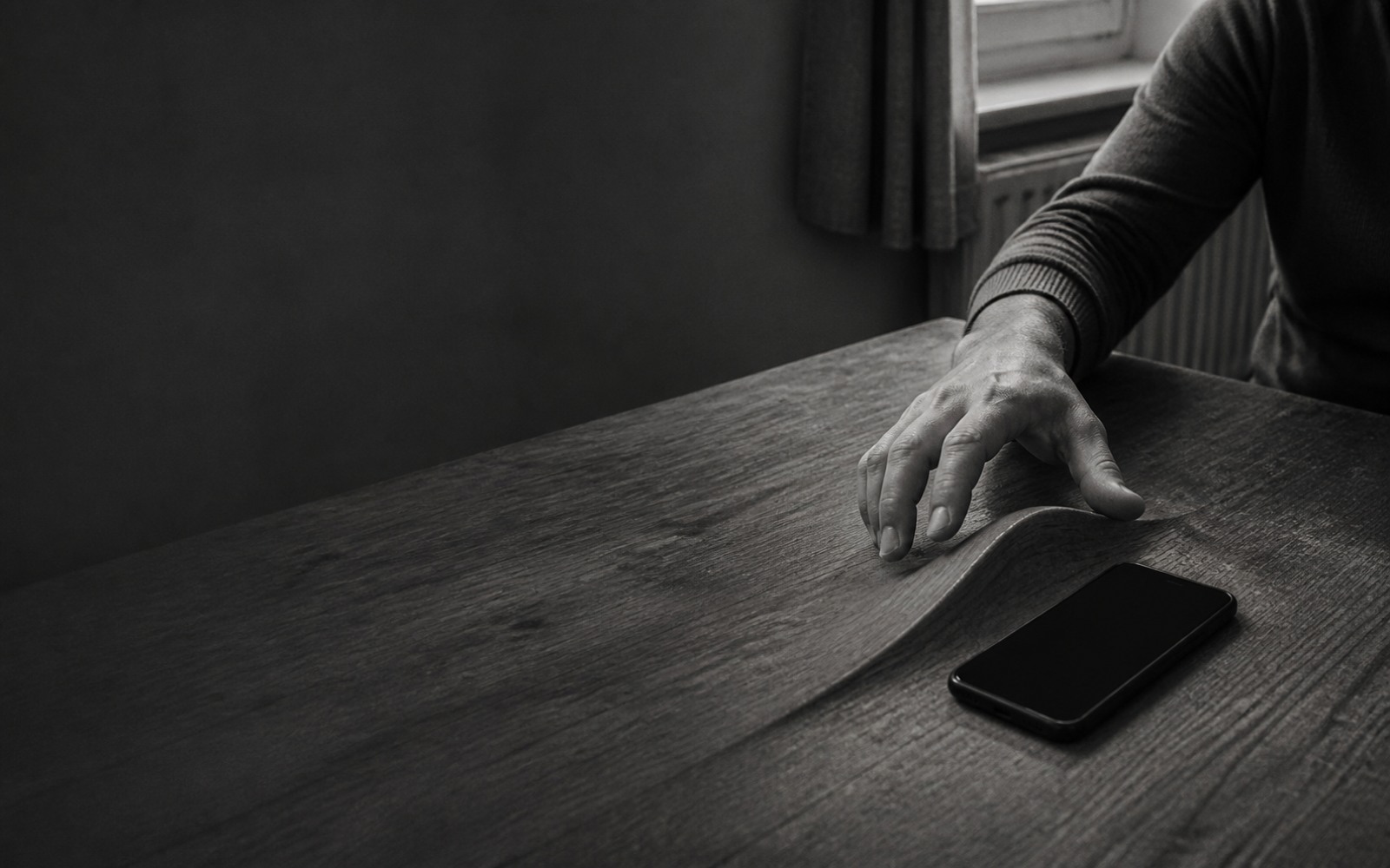 A hand rests on a wooden table, fingertips at a raised fold in the tabletop; beyond the fold lies a phone with a blank screen.