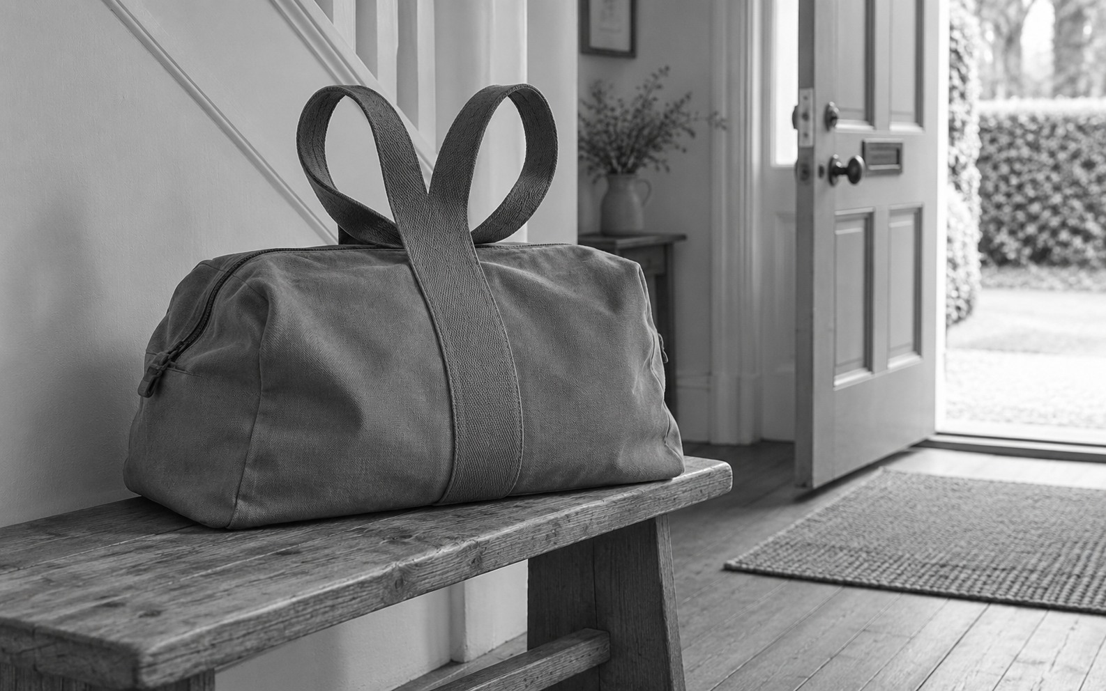 A canvas bag rests on a hallway bench with two upright loop handles branching from a broad central strap.