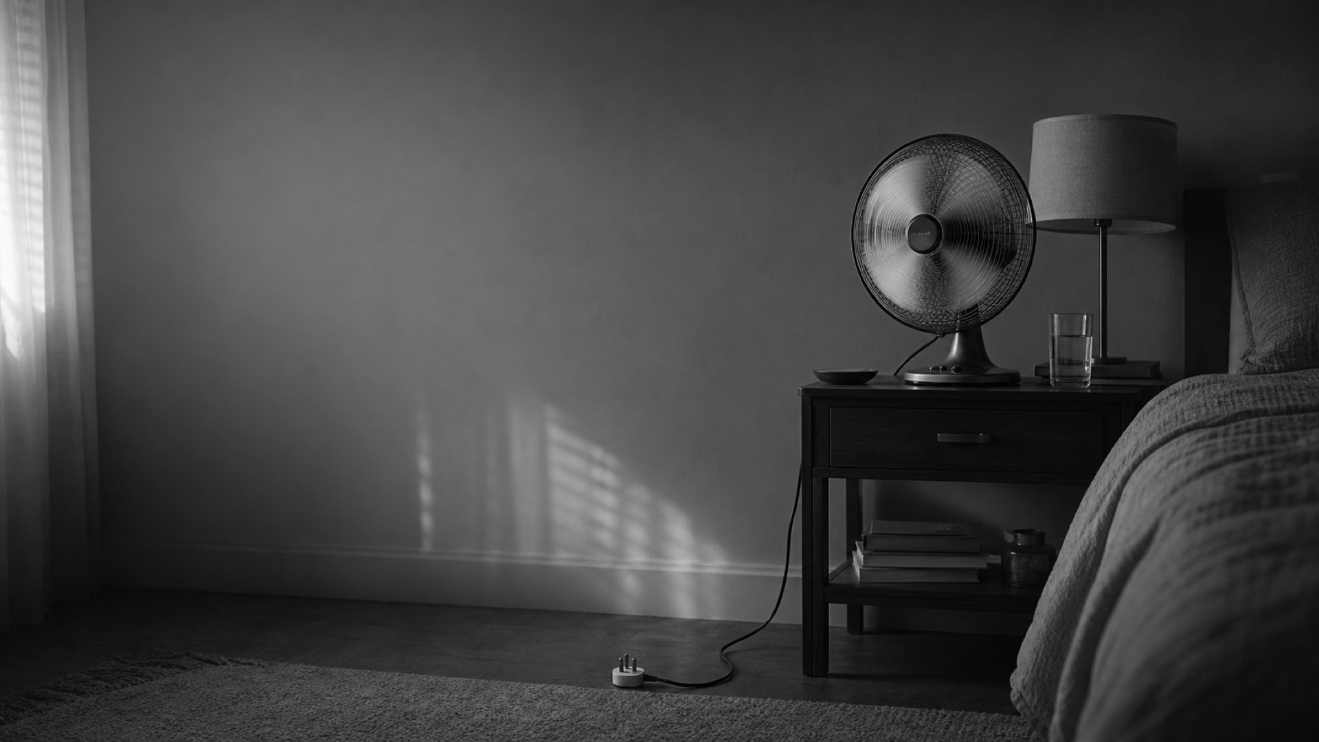An empty bedroom at dusk. On the bedside table a desk fan turns beside a lamp and a glass of water, its cord trailing to the socket.