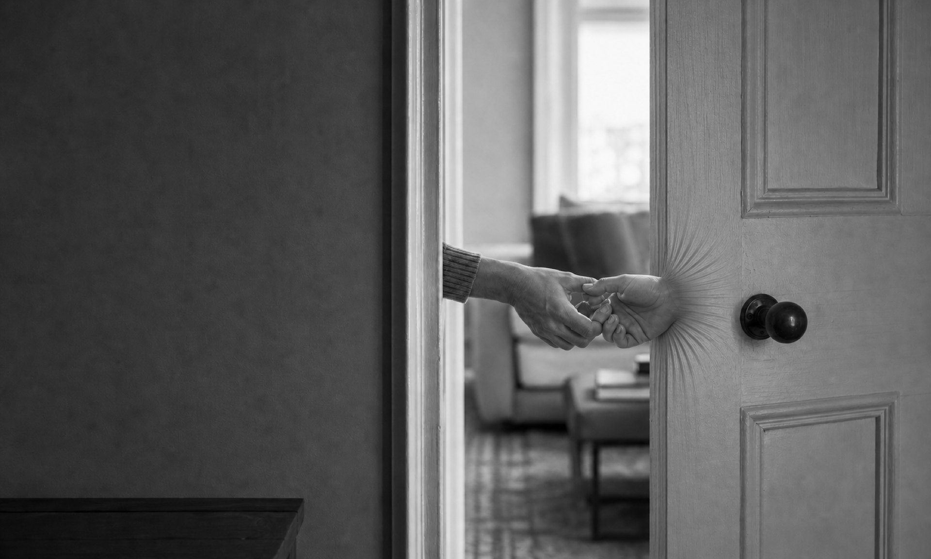 A door stands part open onto a lit sitting room. A sleeved arm reaches out through the gap, and from the door itself a second hand emerges, the grain of the wood radiating around its wrist, the two hands clasped.
