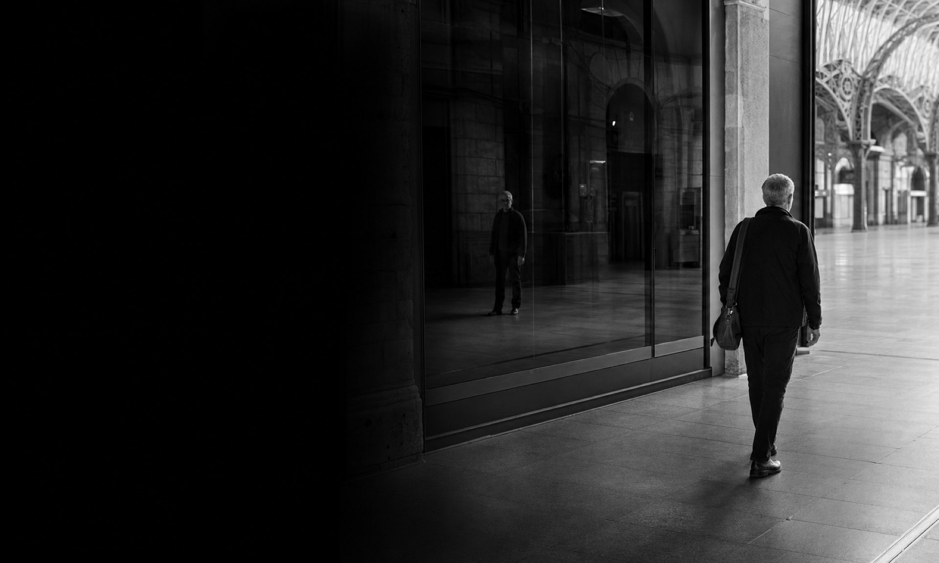 A man with a bag walks along the glass front of a station concourse. His reflection in the glass stands still, facing the other way.
