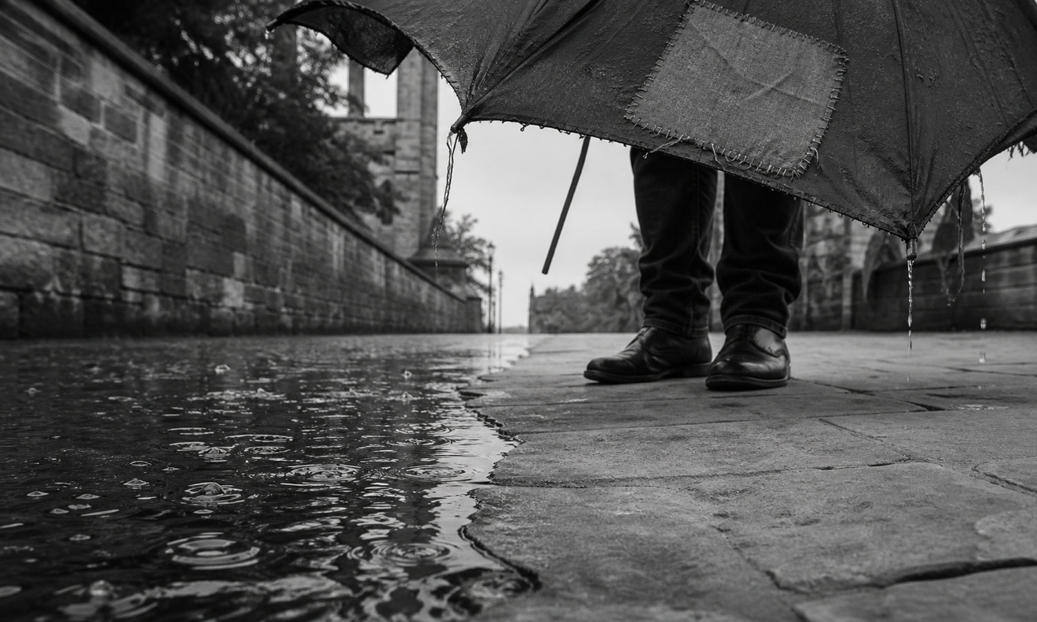 Rainwater ripples across a pavement until it reaches the dry stone beneath a lone person holding a torn, patch-repaired umbrella with a broken rib.