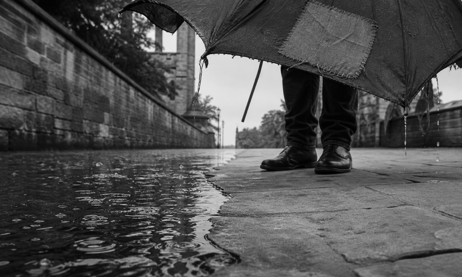 Rainwater ripples across a pavement until it reaches the dry stone beneath a lone person holding a torn, patch-repaired umbrella with a broken rib.