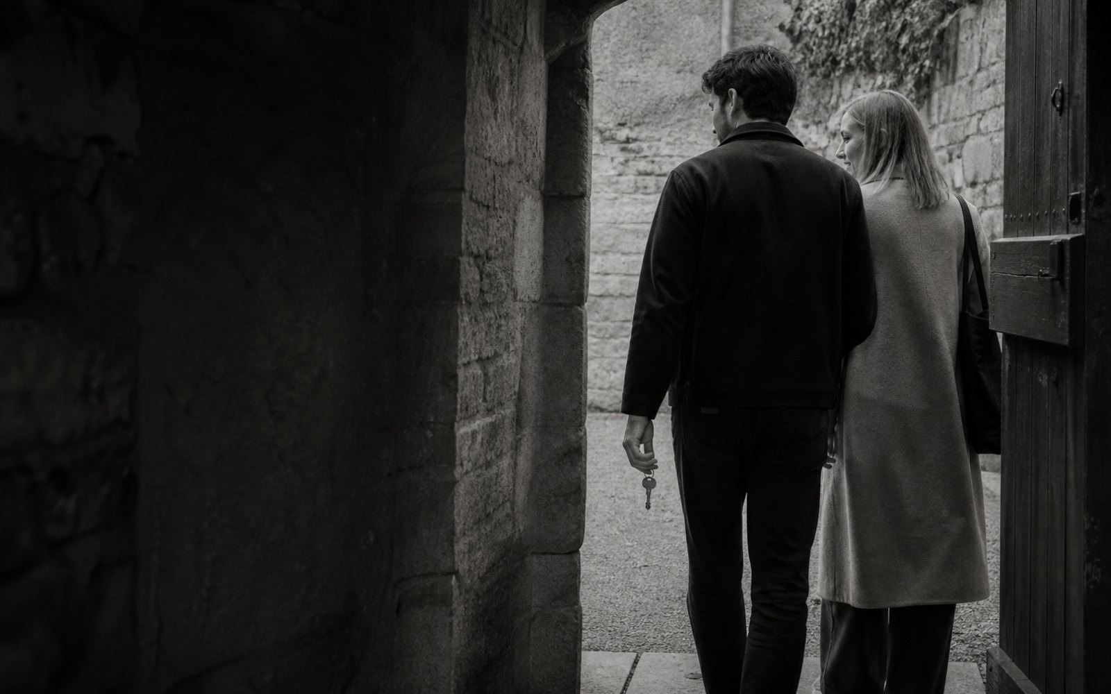 A younger man carries an ordinary metal key beside a fair-haired woman as they approach an open, unheld gate.