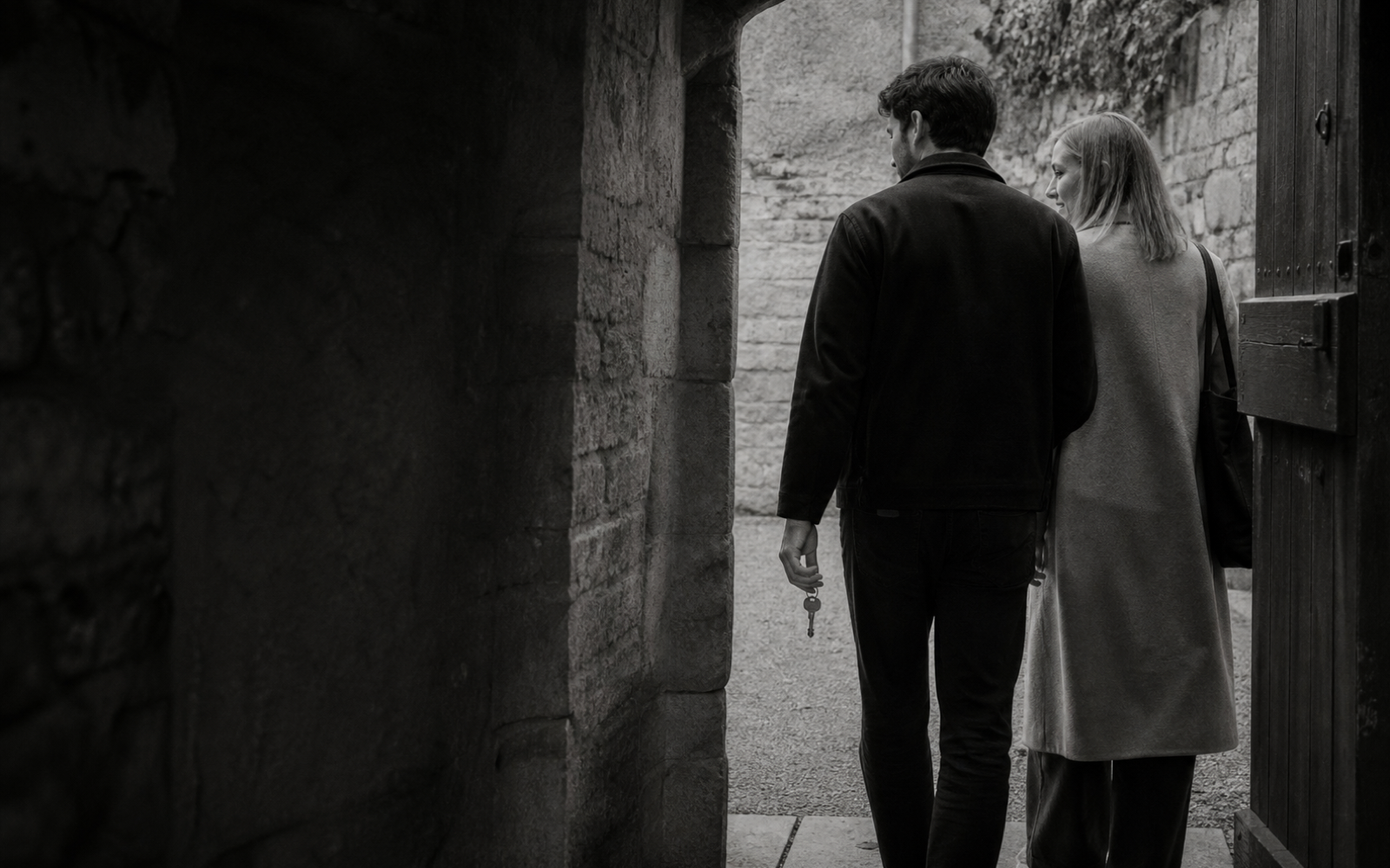 A younger man carries an ordinary metal key beside a fair-haired woman as they approach an open, unheld gate.