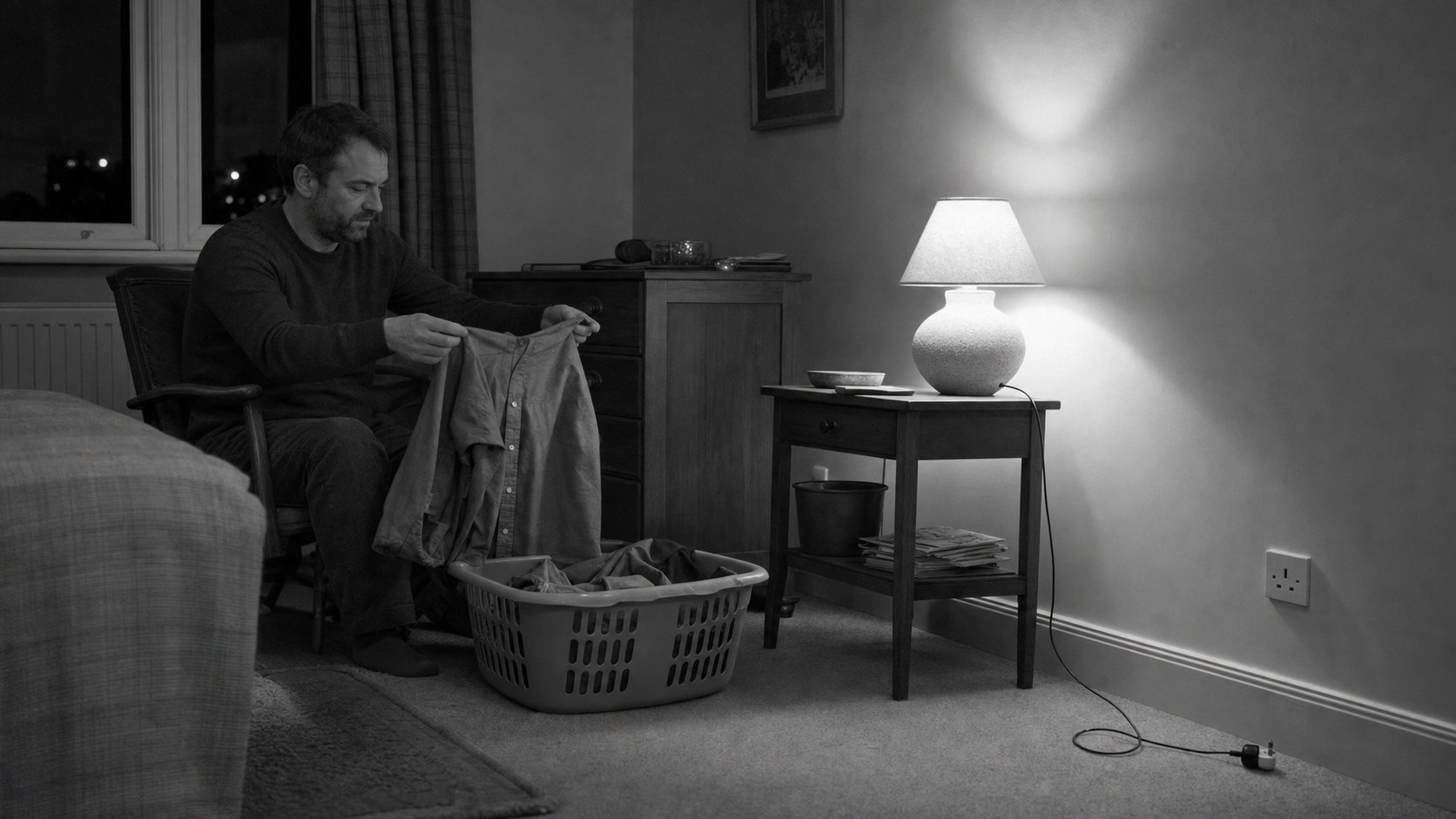 A man sits alone on the edge of a bed at night, folding laundry by lamplight while the rest of the room stays dark.