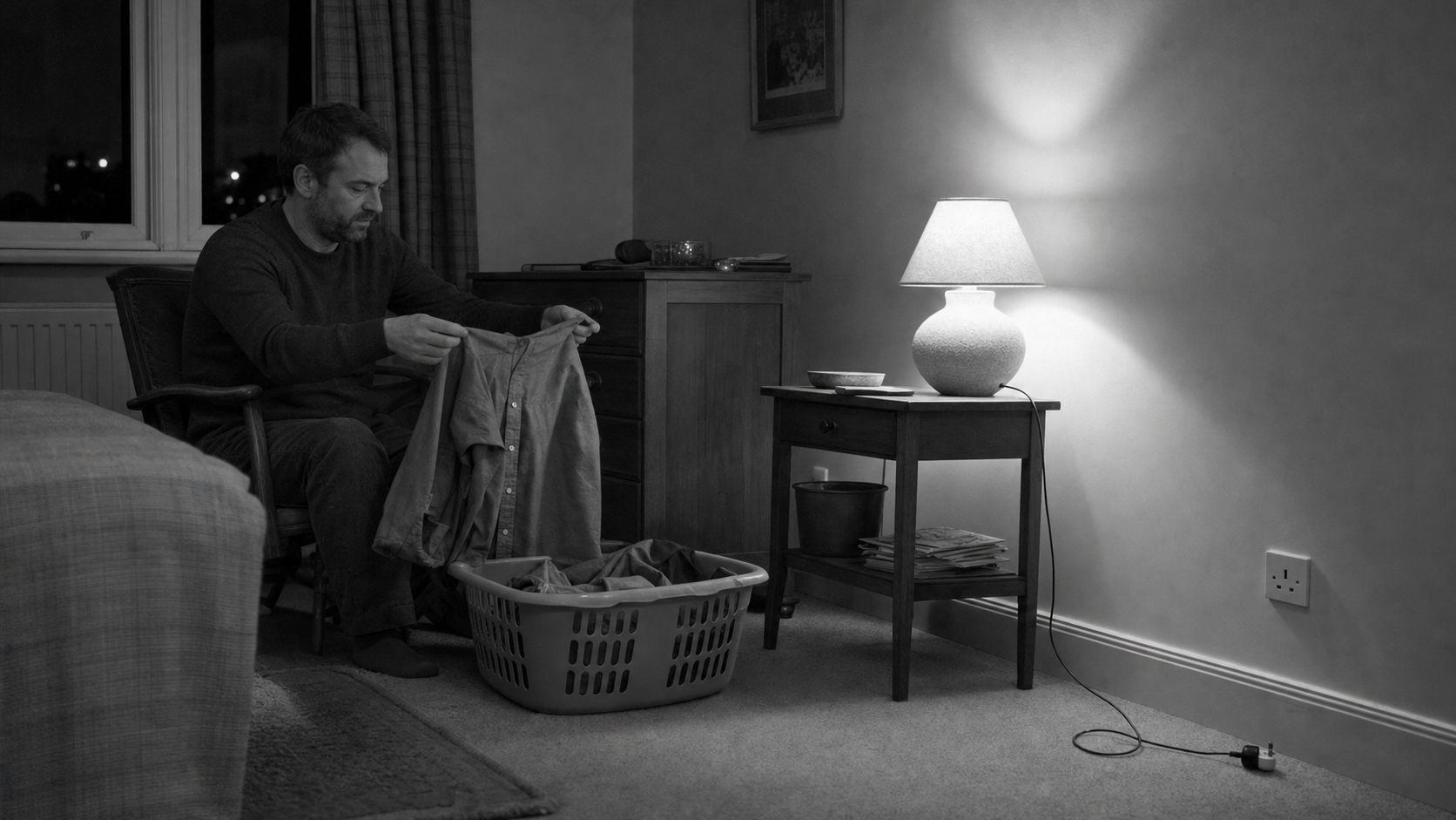A man sits alone on the edge of a bed at night, folding laundry by lamplight while the rest of the room stays dark.
