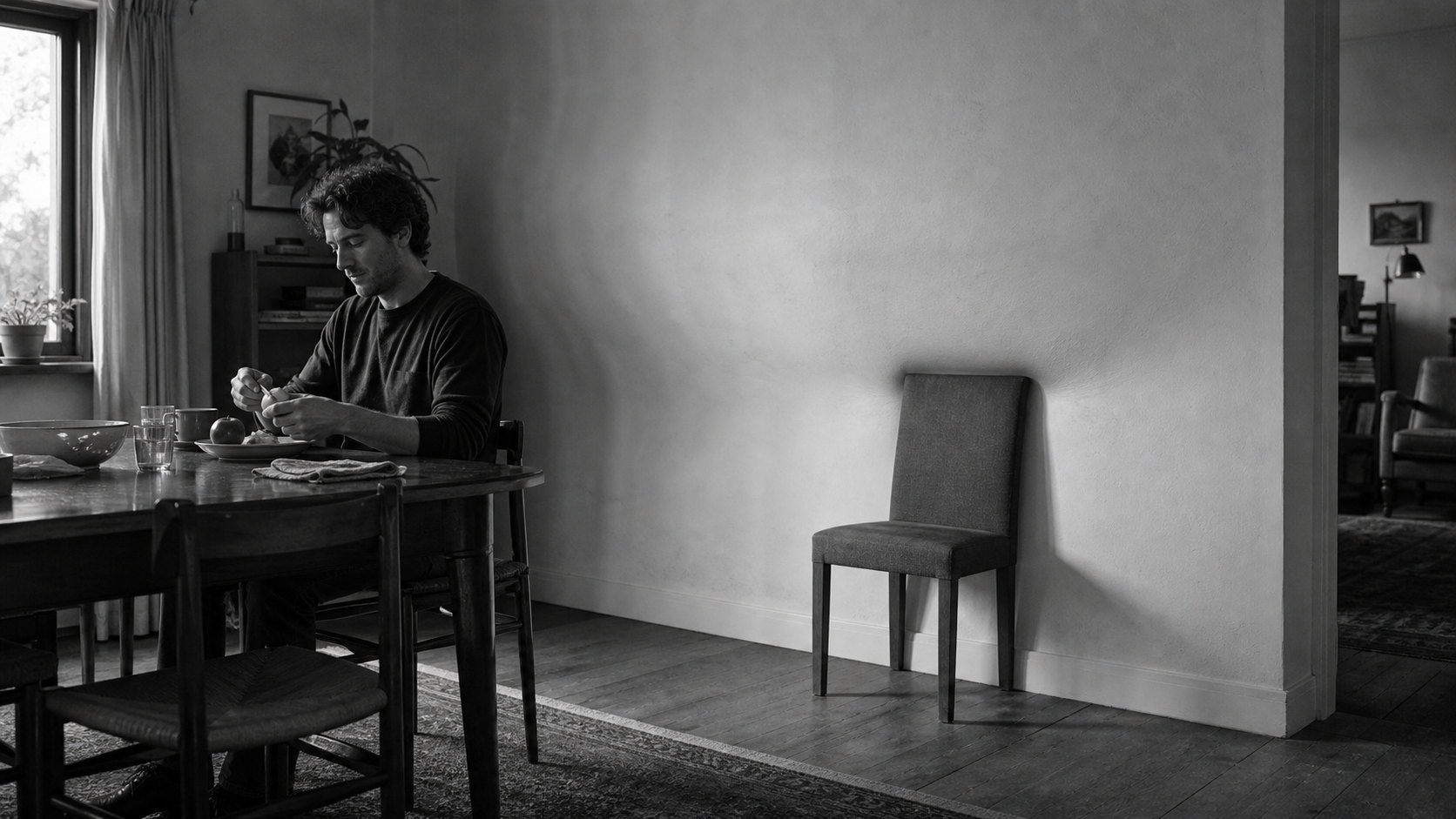 A man eats alone at a dining table while a single empty chair stands apart from the others, lit against a bare wall.