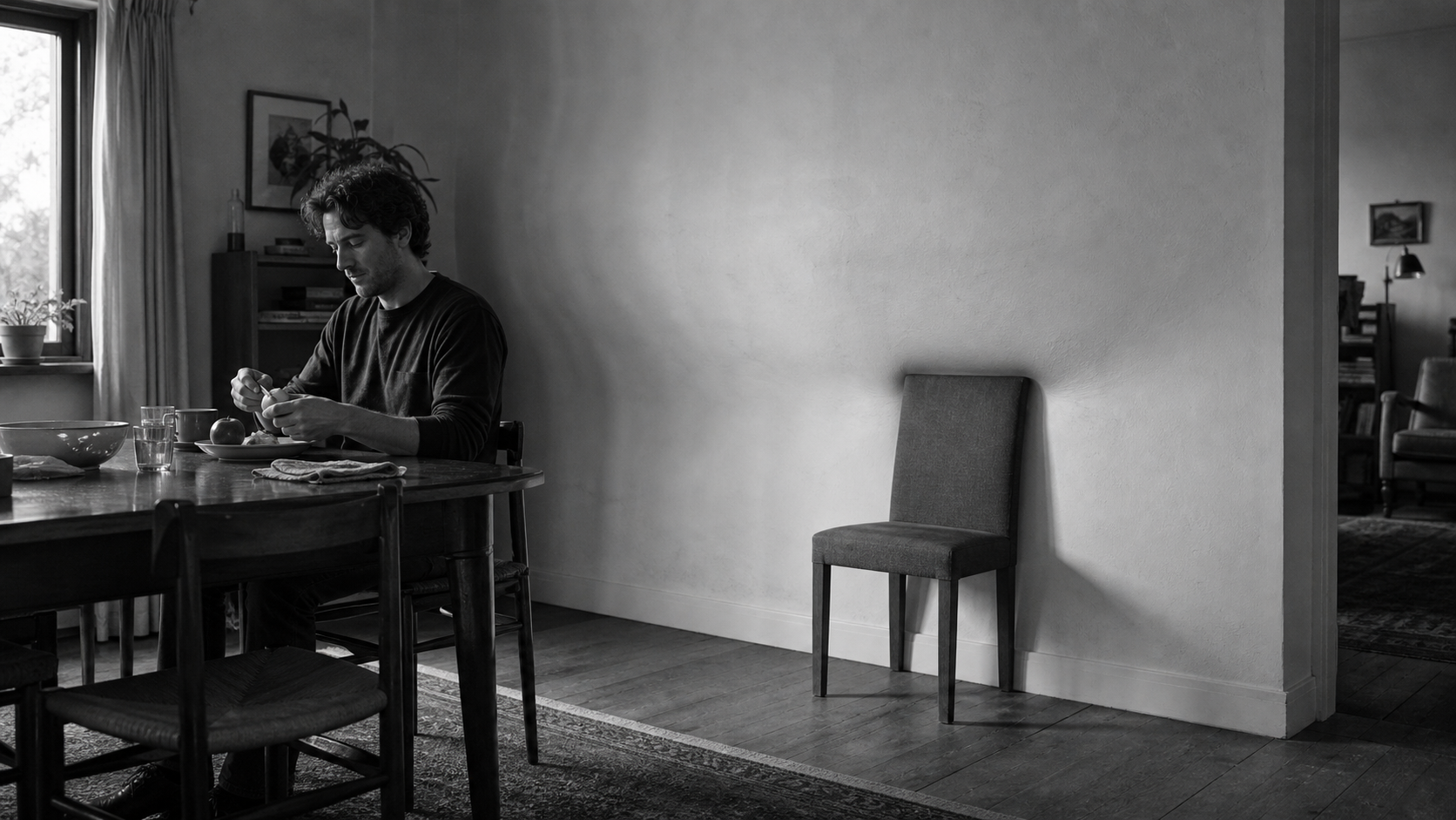 A man eats alone at a dining table while a single empty chair stands apart from the others, lit against a bare wall.