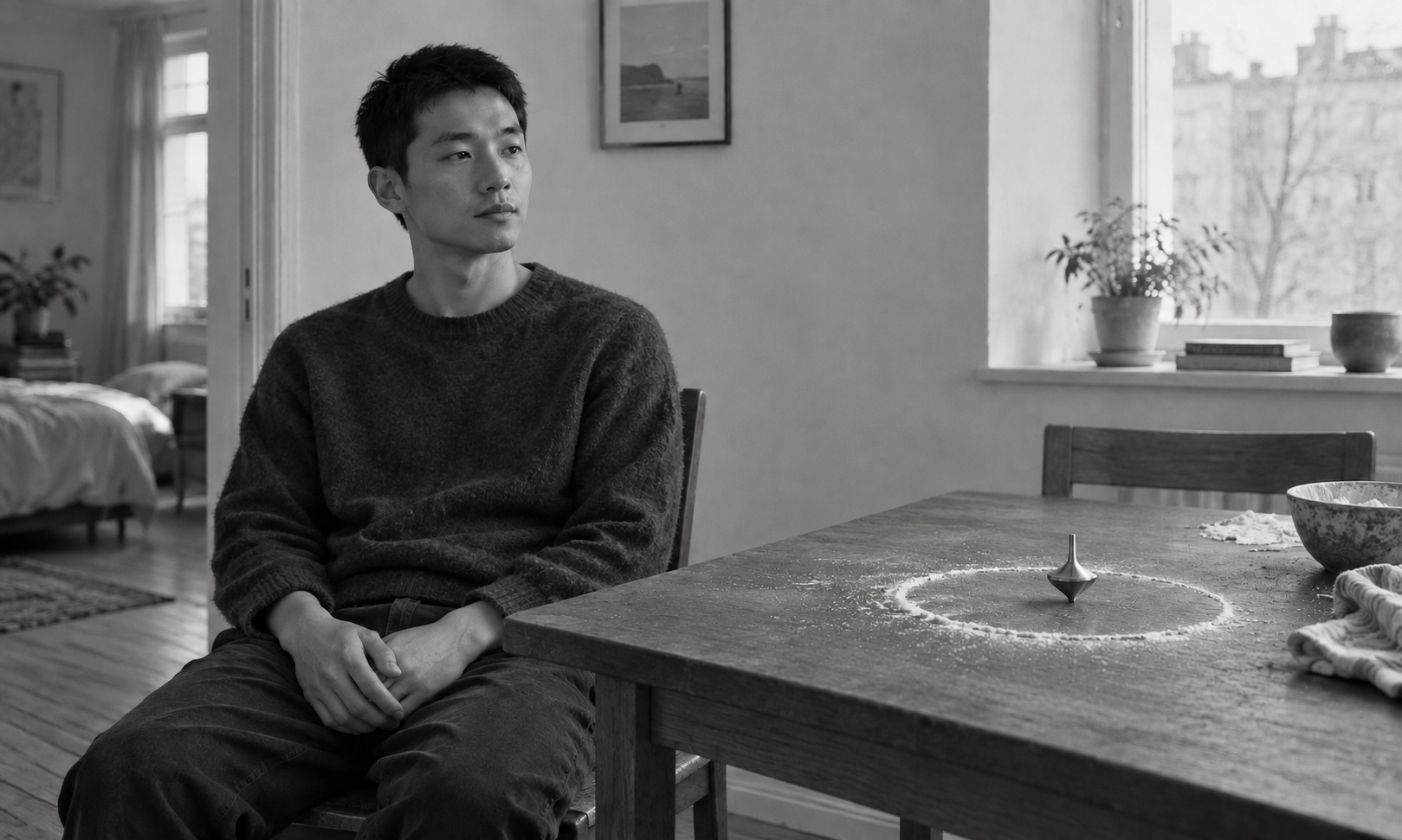 A man sits at a wooden table, hands in his lap. On the table a small metal spinning top stands upright inside a circle of flour.