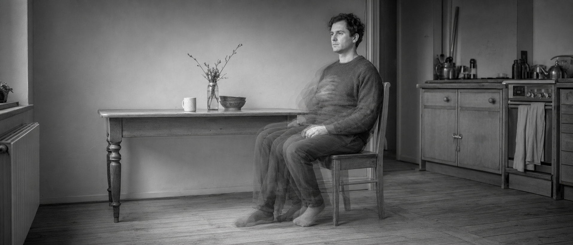 A man sits still at a kitchen table while his body blurs with movement beneath a perfectly sharp head.