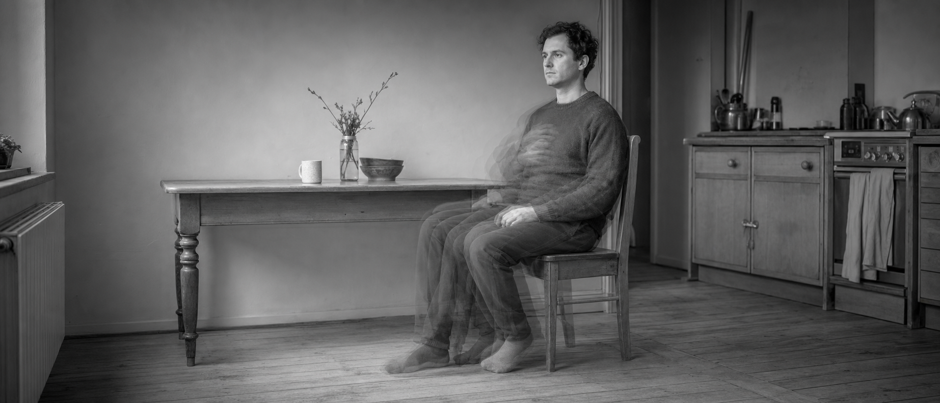 A man sits still at a kitchen table while his body blurs with movement beneath a perfectly sharp head.