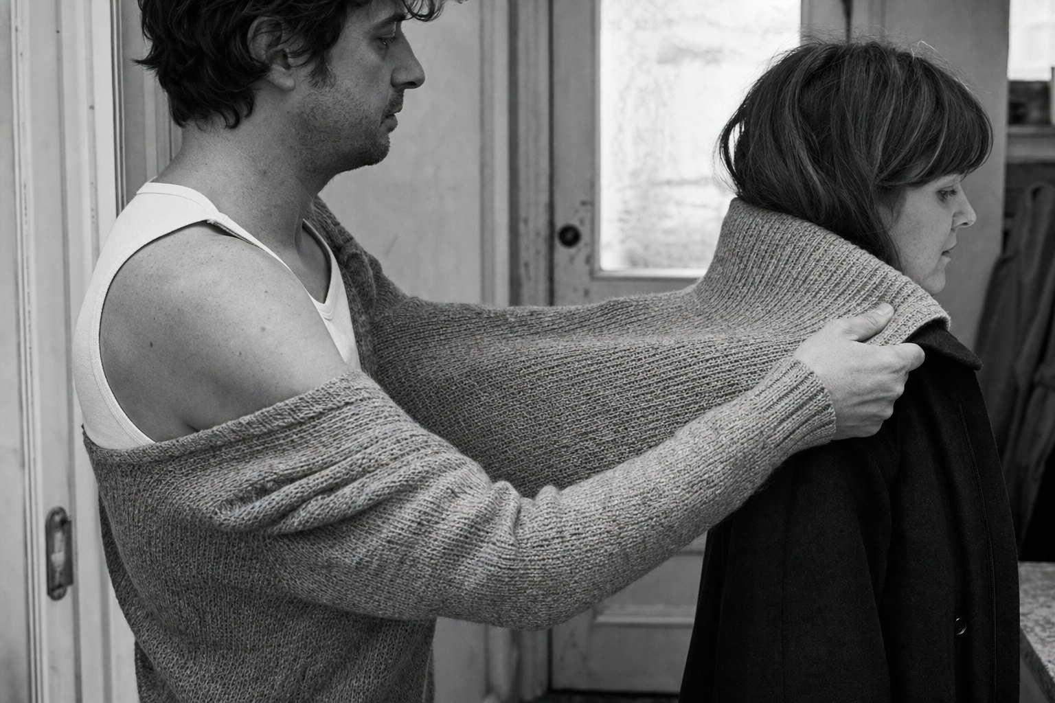 A man lifts his own jumper off his back and drapes it around a woman in a coat, leaving himself in a thin vest.