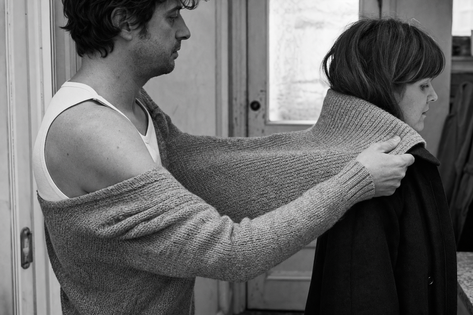 A man lifts his own jumper off his back and drapes it around a woman in a coat, leaving himself in a thin vest.