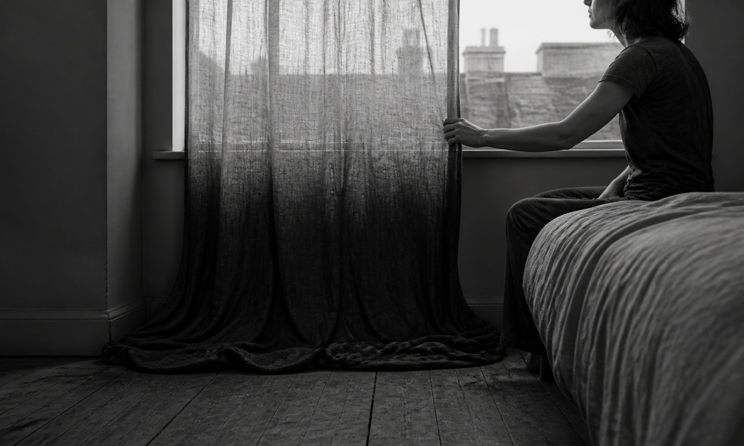 A person sits beside a curtain whose dense pooled hem rises into sheer fabric at the morning window.