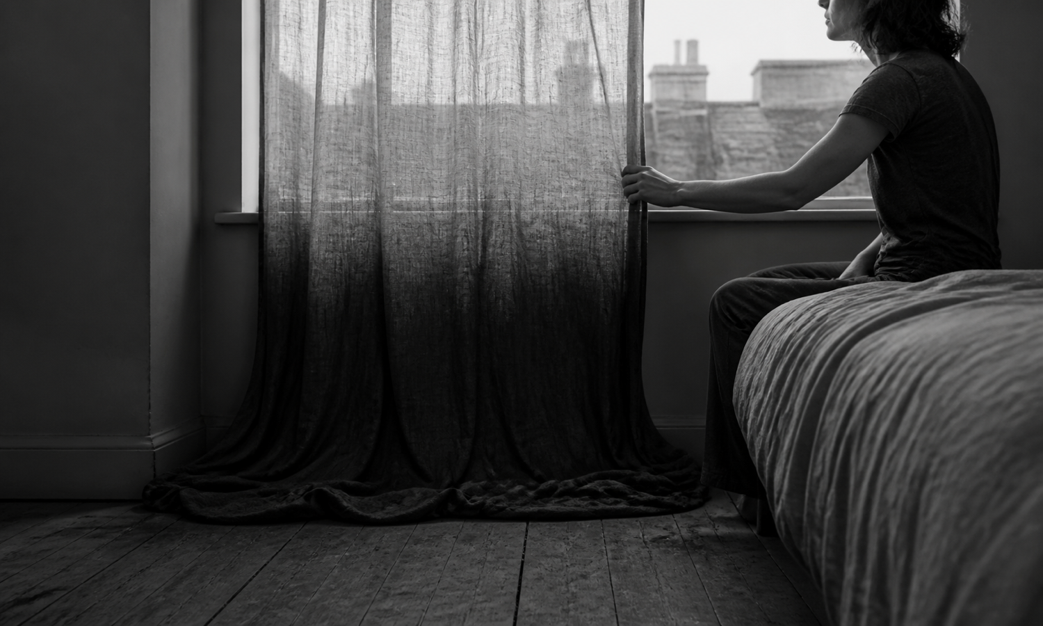 A person sits beside a curtain whose dense pooled hem rises into sheer fabric at the morning window.