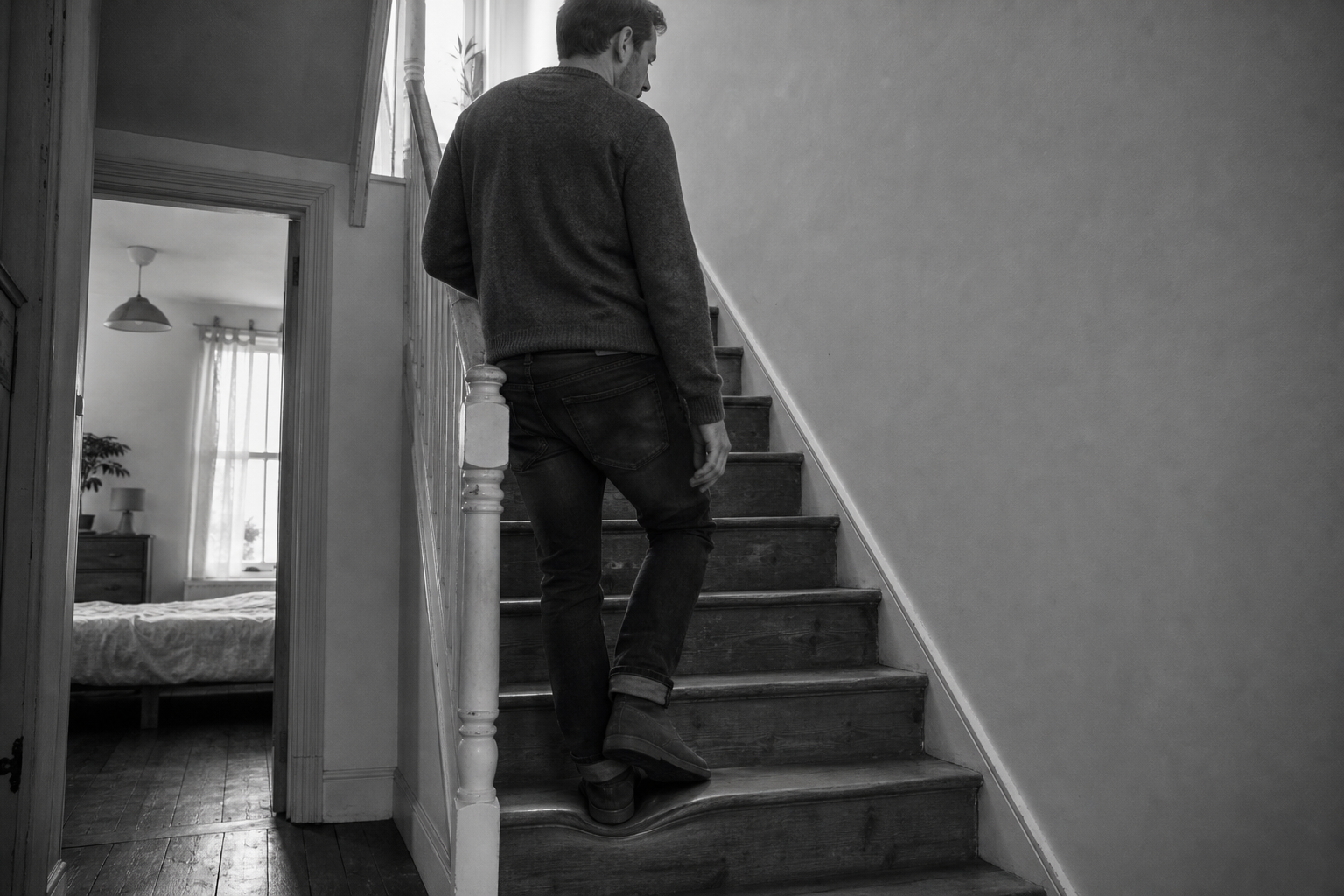 A man pauses partway up a staircase, one hand on the wall, having stopped between the bedroom below and the landing above.