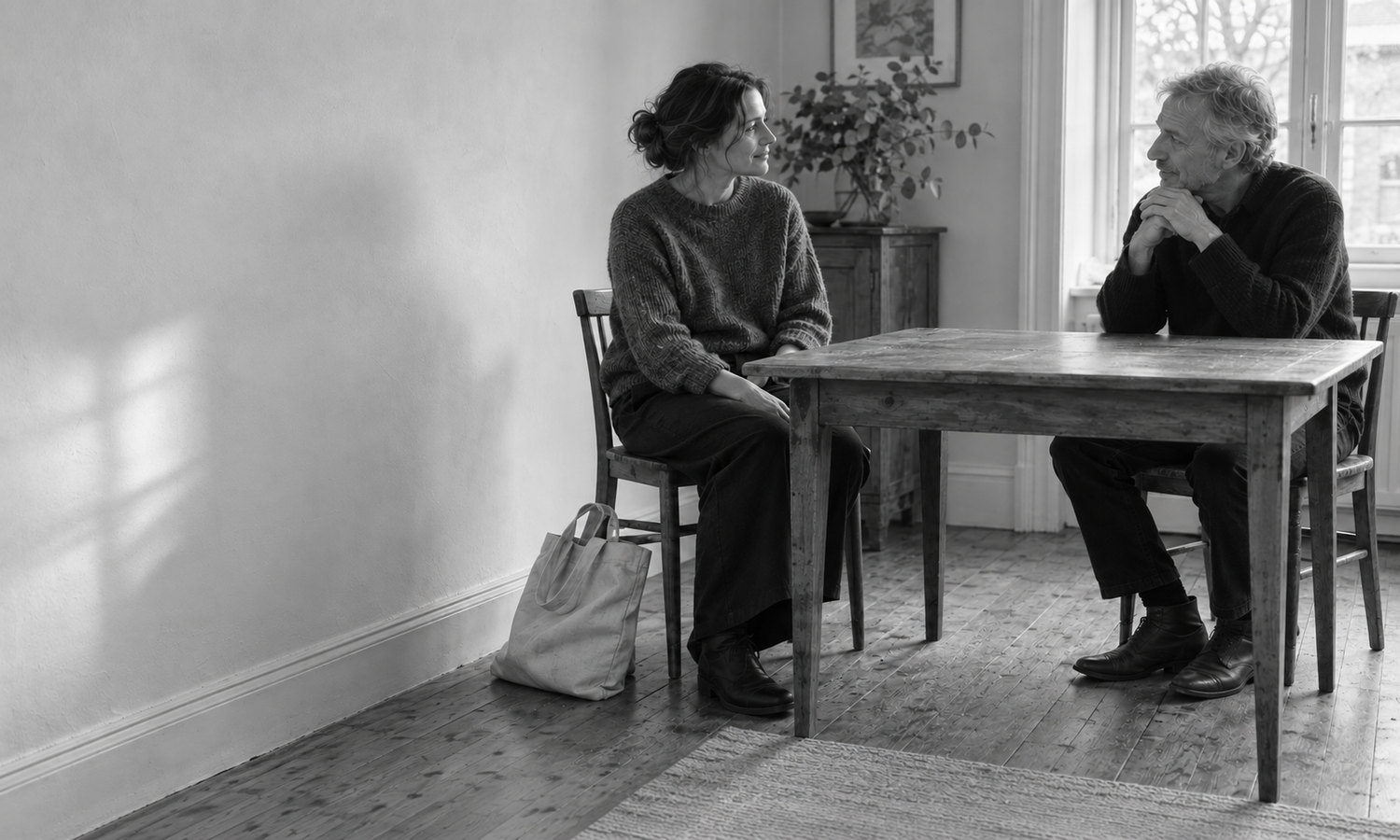 A woman and an older man sit facing one another across a wooden table, with a bag beside the woman's chair.