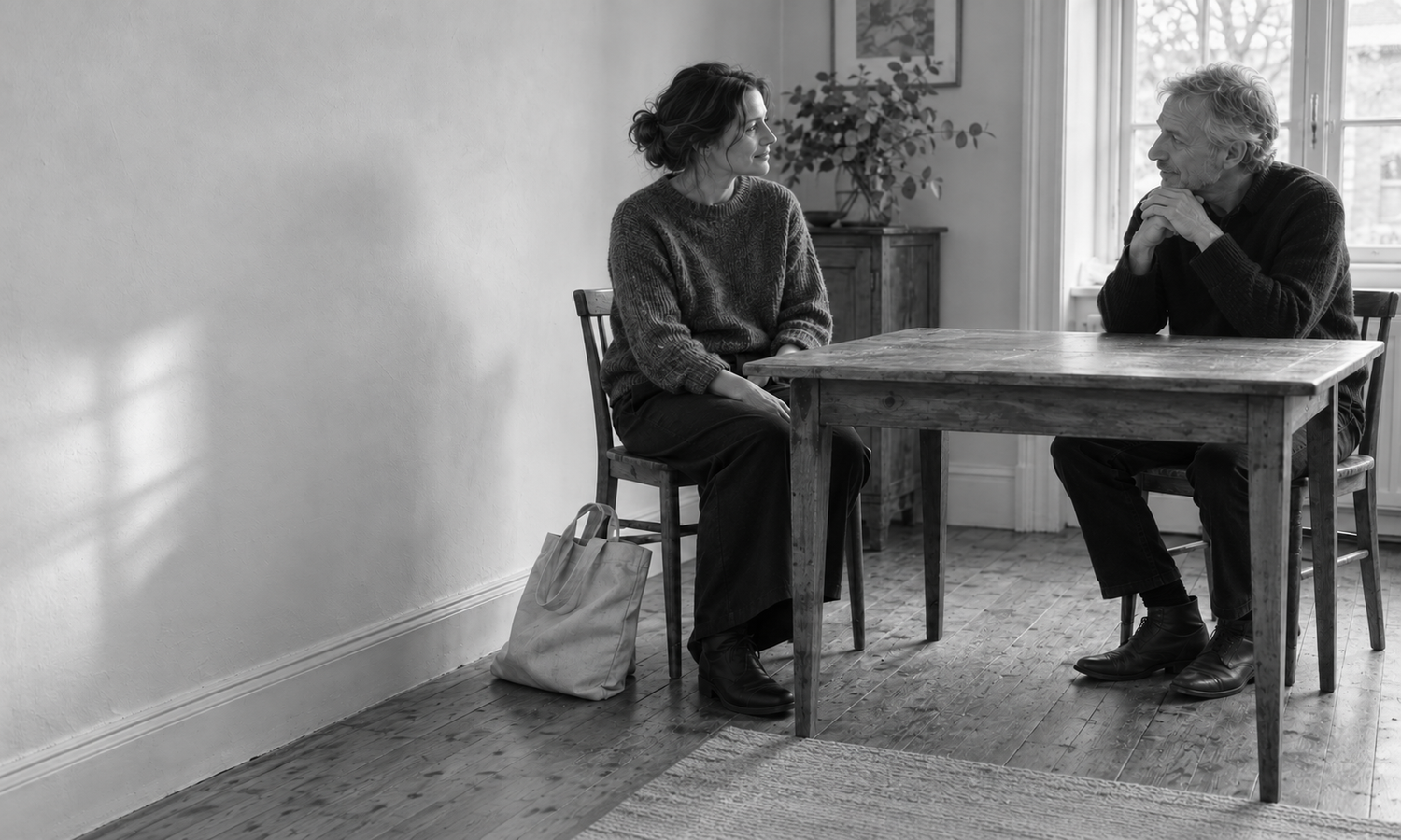 A woman and an older man sit facing one another across a wooden table, with a bag beside the woman's chair.