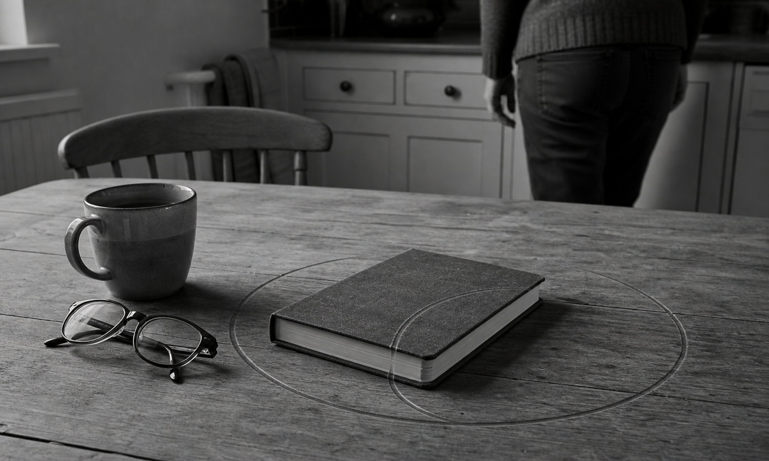 A closed clothbound notebook lies on a wooden kitchen table as a circular wear groove continues from the wood across its cover and paper edges while an adult walks away.