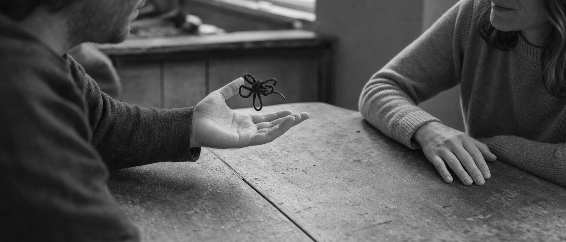 Two people sit across a table; one holds out an open palm with a small dark knot resting in it, while the other keeps both hands flat on the wood.