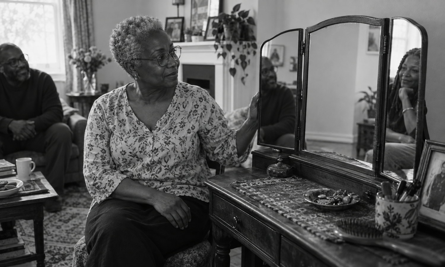A grey-haired woman in glasses holds the edge of a three-panel mirror; its side panes reflect two seated people while the central pane shows a pale wall and floor.