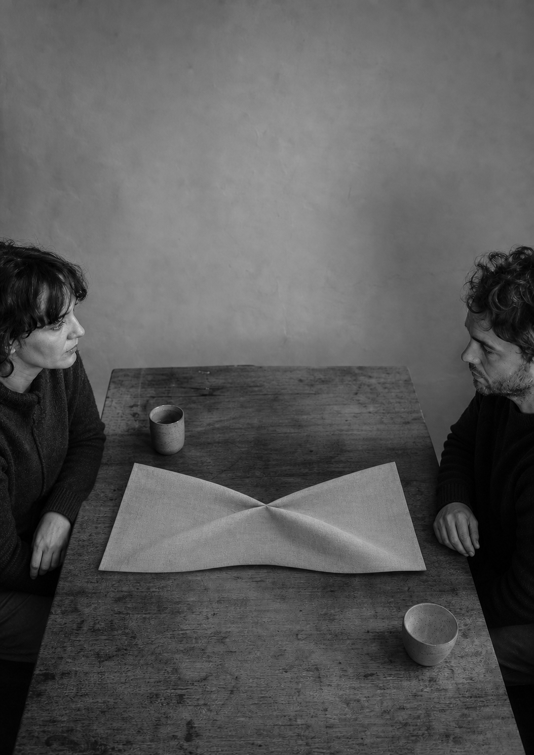 Two people sit at opposite ends of a table over a sheet of paper folded so that both halves rise away from the middle.