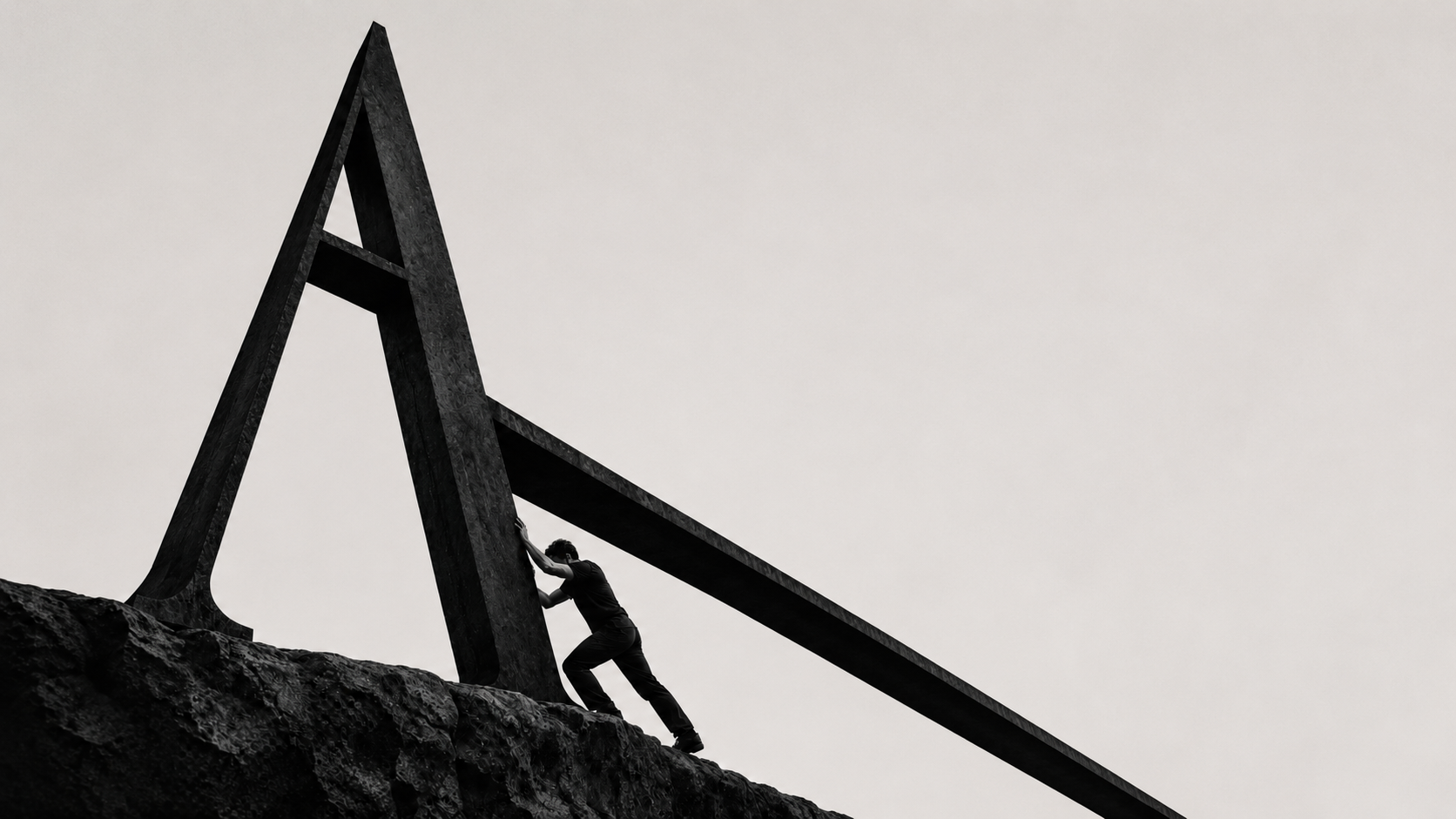A lone figure straining to push a monumental serif letter ‘A’ upright on a rocky ledge against a pale sky.