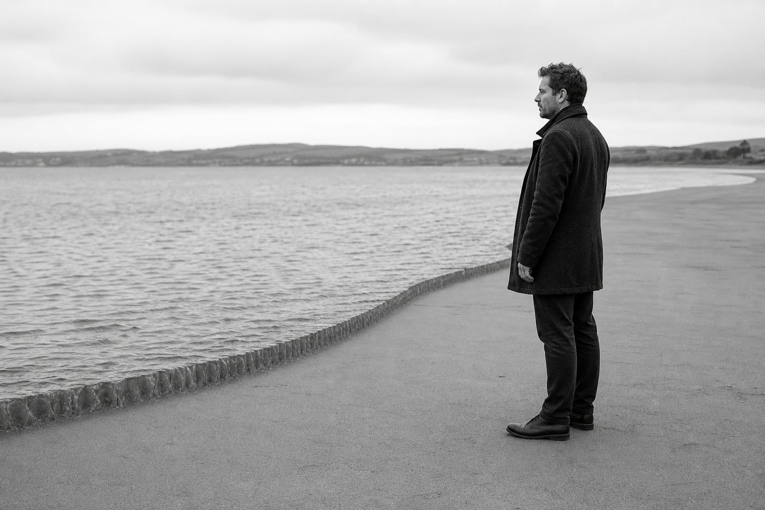 A man stands at the edge of a wide shore watching the water, the tideline drawn out flat in front of him.