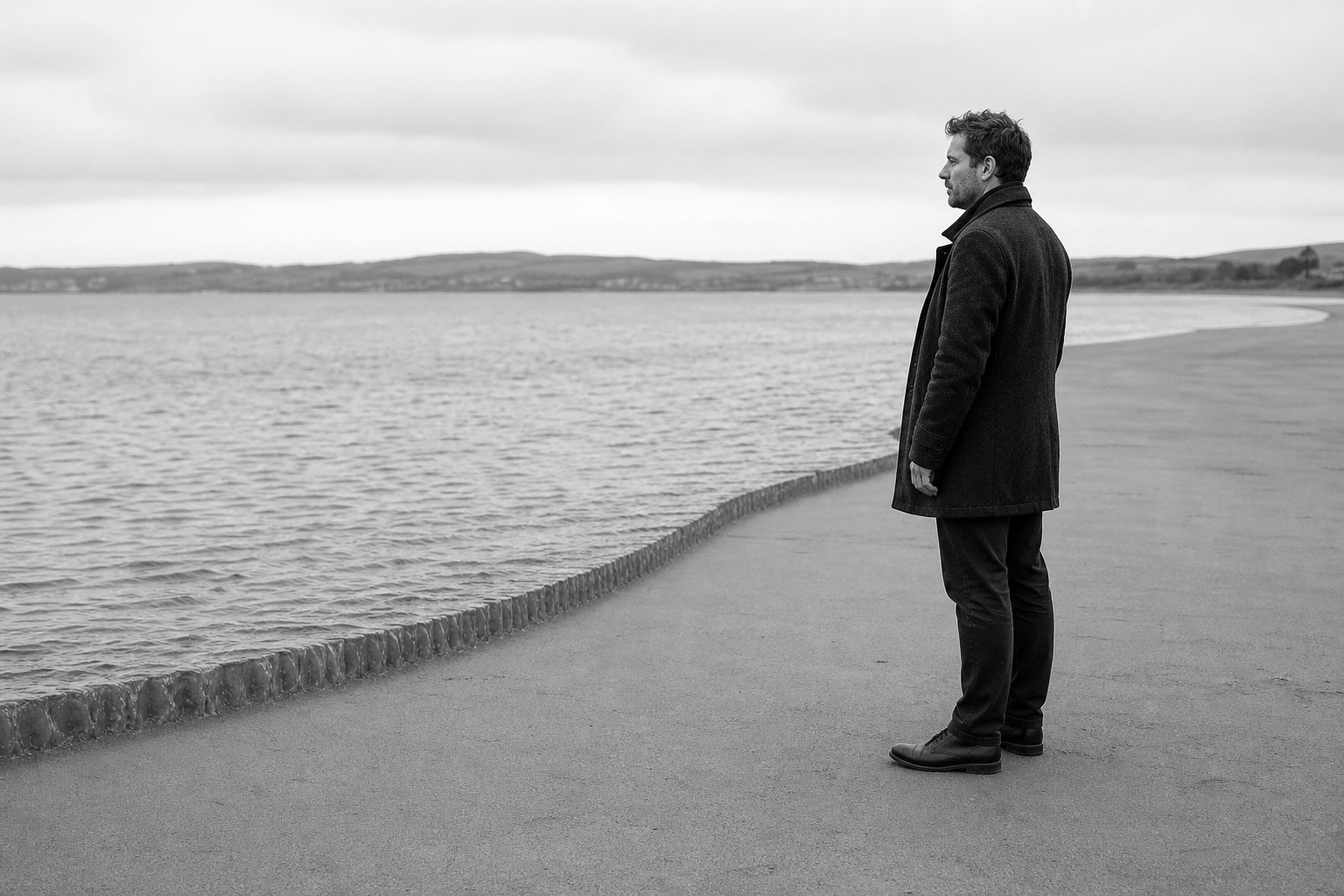 A man stands at the edge of a wide shore watching the water, the tideline drawn out flat in front of him.