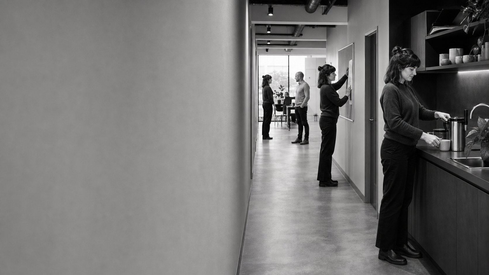 A woman pours coffee at the end of a workplace corridor while colleagues talk and pin up a notice further down.