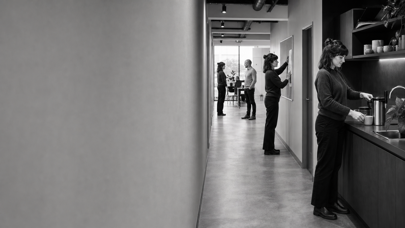 A woman pours coffee at the end of a workplace corridor while colleagues talk and pin up a notice further down.