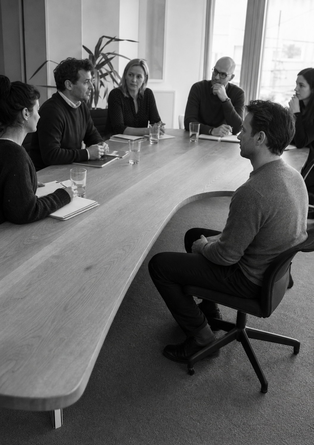 A meeting continues along a boardroom table that swells and buckles beside one seated man, leaving him below the line of the others.