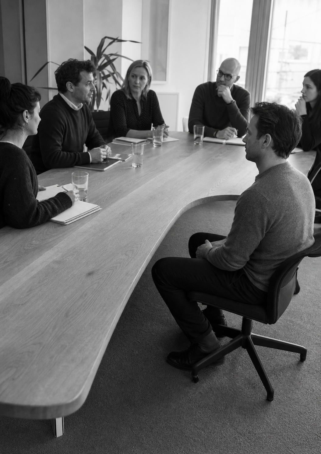A meeting continues along a boardroom table that swells and buckles beside one seated man, leaving him below the line of the others.