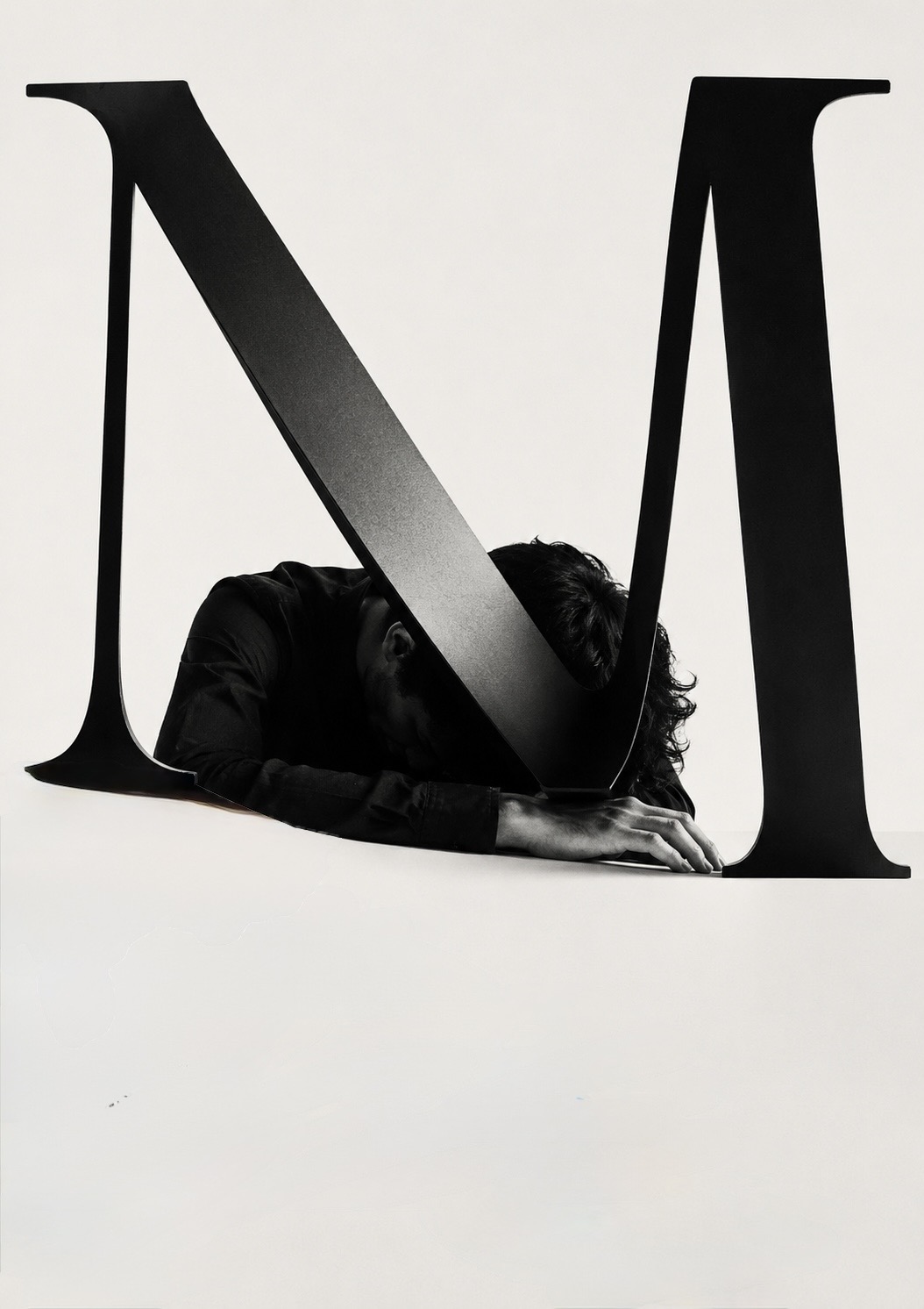 A large sculpted letter M with a person slumped face-down beneath its strokes, arms outstretched on the surface.