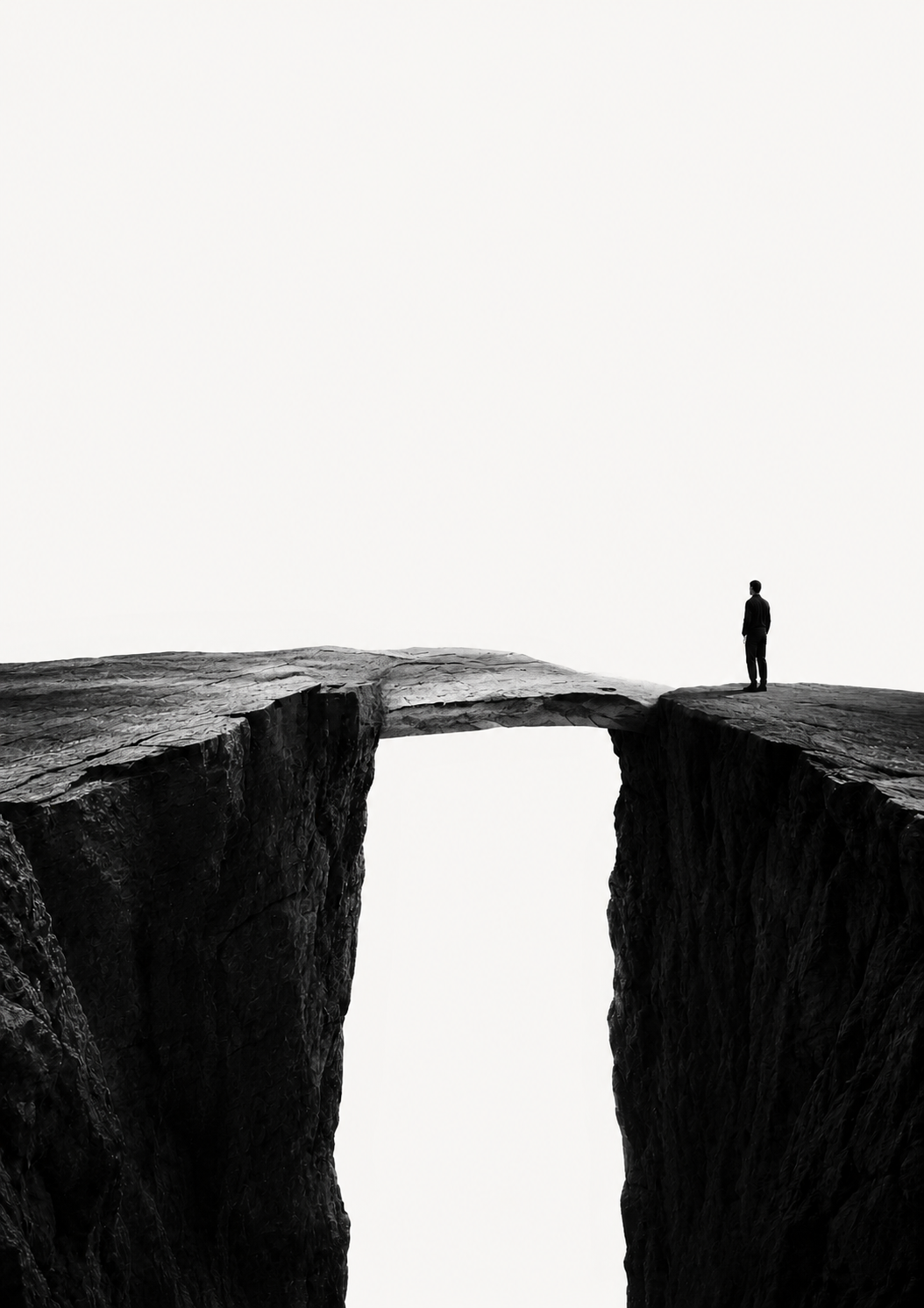 A lone figure standing at the right edge of a vast dark chasm that is spanned by a single natural rock bridge, under a pale open sky.