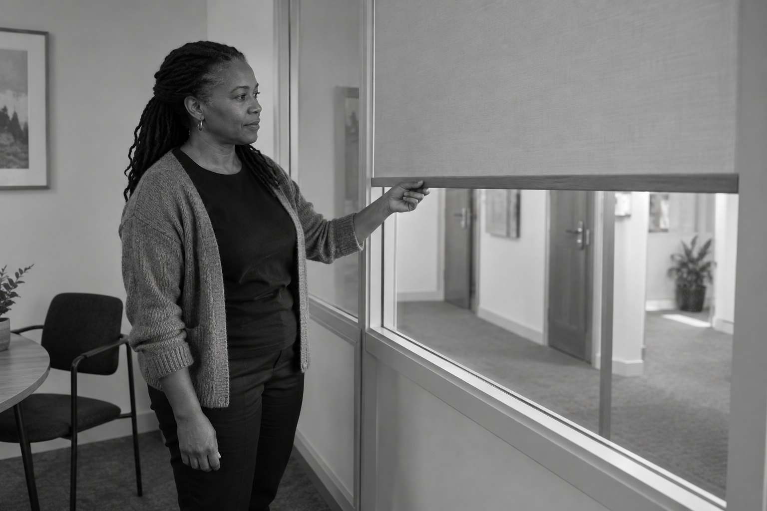 A woman holds the wooden bottom rail of a partly lowered fabric blind across an internal office window, with an empty corridor visible through the uncovered lower glass.